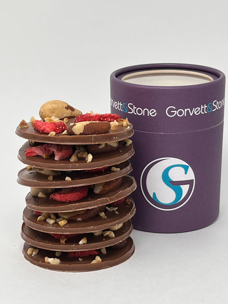 Stack of large milk chocolate mendiant discs topped with dried fruits and nuts with the box behind. On a white background.