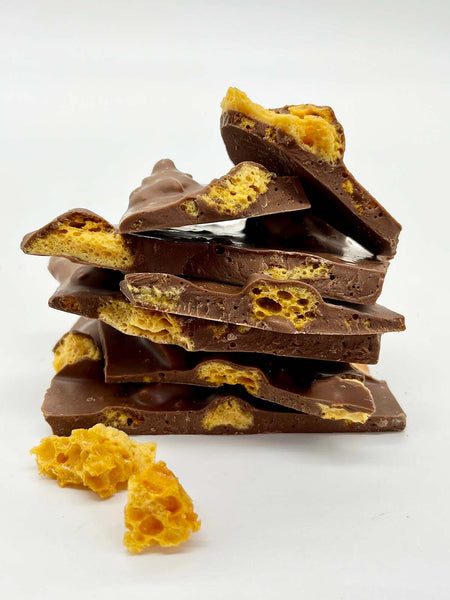Stack of handmade milk chocolate honeycomb shards with 2 small chunks of naked honeycomb in front. View taken at eye level on a white background.