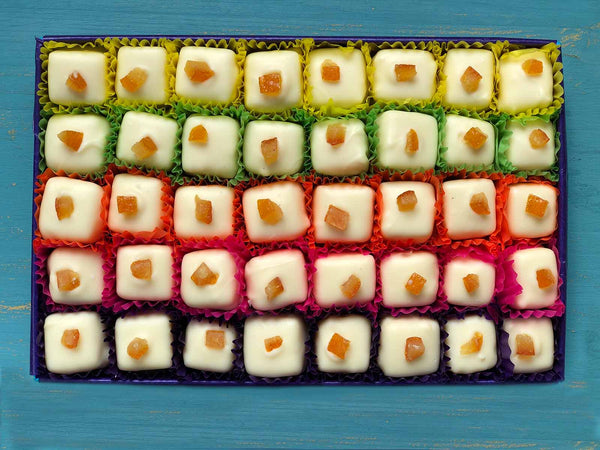 Open gift box of 40 white chocolates decorated with candied orange pieces. Each chocolate is contained in a colourful petits fours case. View from above on a painted blue background.