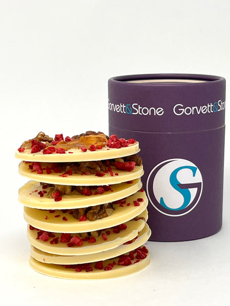 Stack of giant white chocolate mendiants topped with rasberries, mango and walnuts with the box behind. On a white background