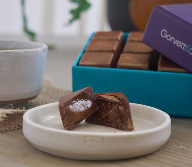 One sticky toffee caramel chocolate on a plate cut open revealing the caramel inside. An open box of the chocolates in the background. 