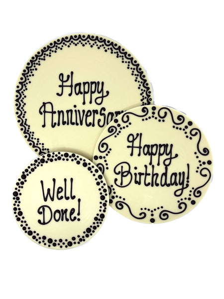 3 White chocolate plaques hand piped with decorative borders and 'Happy Anniversary' 'Happy Birthday!' and 'Well Done!' text in dark chocolate. On a white background.
