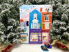 A colorful chocolate advent calendar depicting buildings and shops with Christmas decorations, placed on a table with a Christmas tree in the background.