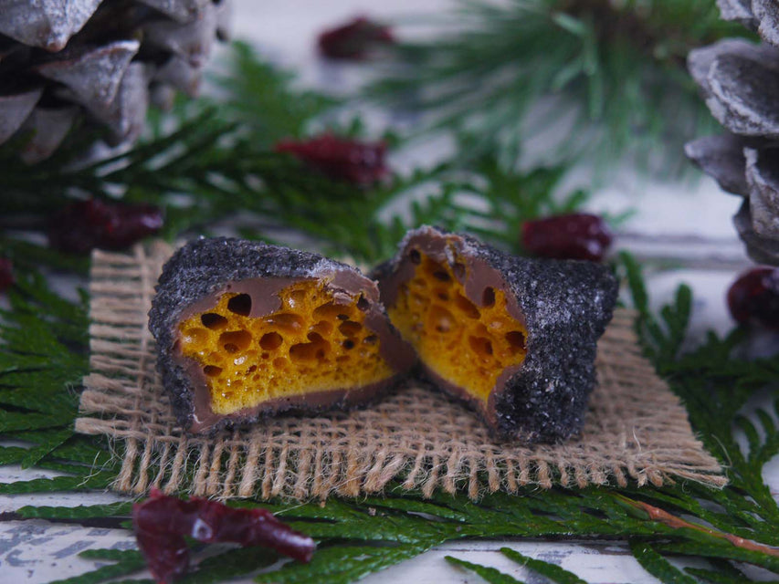 Chunk of Christmas Coal cinder toffee cut open to reveal the inside.  Product placed on a burlap napkin with greenery and pine cones in the background.
