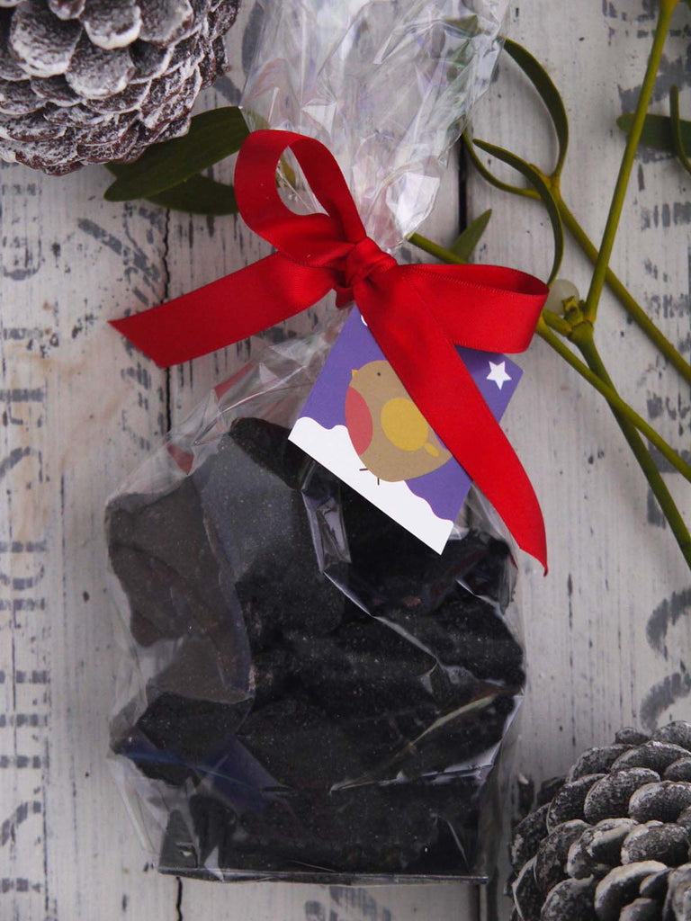 Christmas Coal Cinder Toffee in a clear bag tied with a red ribbon and christmas tag. Viewed from above on a distressed white background.