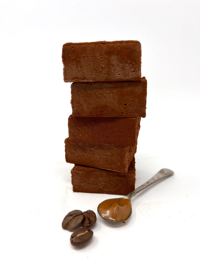 A stack of milk chocolate meltaway squares with coffee beans and a spoon of hazelnut praline on the side. On a white background.