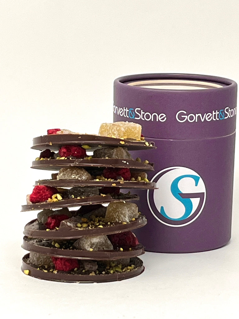 Stack of large dark chocolate mendiant discs topped with dried fruits and nuts with the box behind. On a white background.