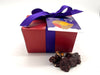 Red box tied with purple ribbon and tag. 2 chocolates in front of the box.  All on a white background.