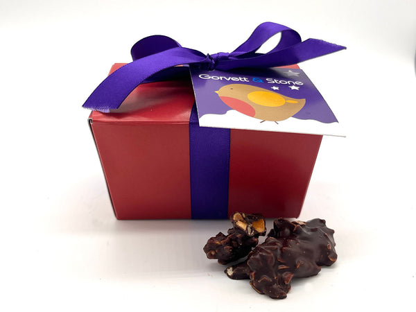 Red box tied with purple ribbon and tag. 2 chocolates in front of the box.  All on a white background.