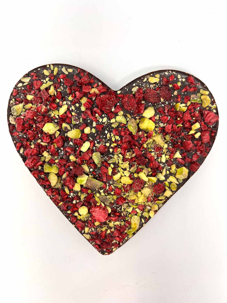 Dark Chocolate Heart decorated with scattered freeze-dried raspberries and crushed pistachios. On a white background.