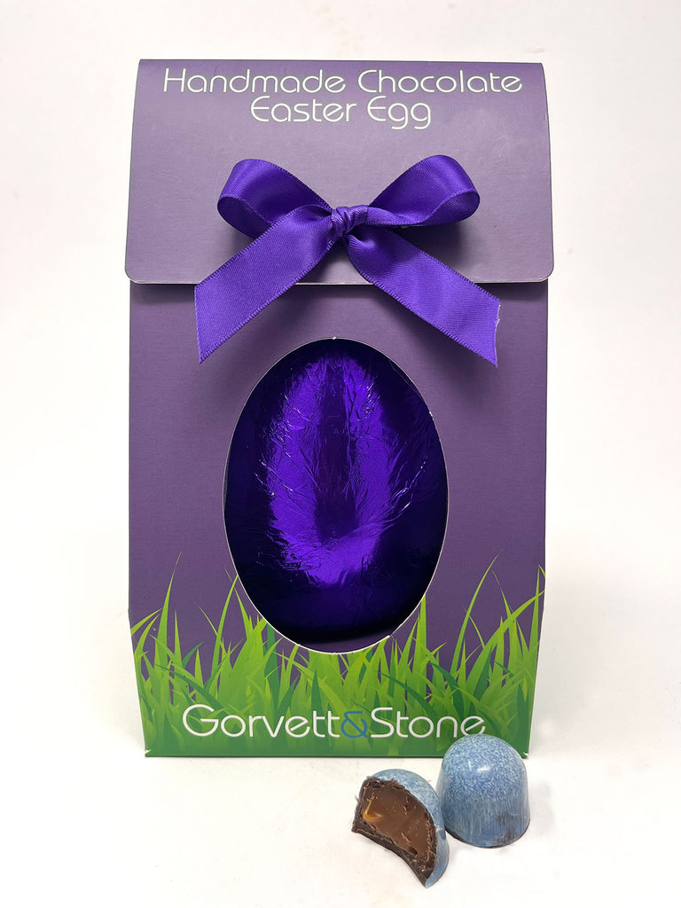 Dark Sea Salt Caramel Easter Egg in Gorvett & Stone Box and purple foil. Two Sea Salt caramel chocolates in the foreground. White background