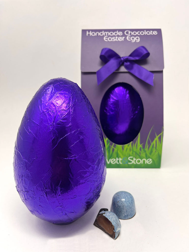 Dark Sea Salt Caramel Easter Egg in purple foil. Gorvett & Stone box in the background and  two Sea Salt Caramel chocolates in the foreground. White background.