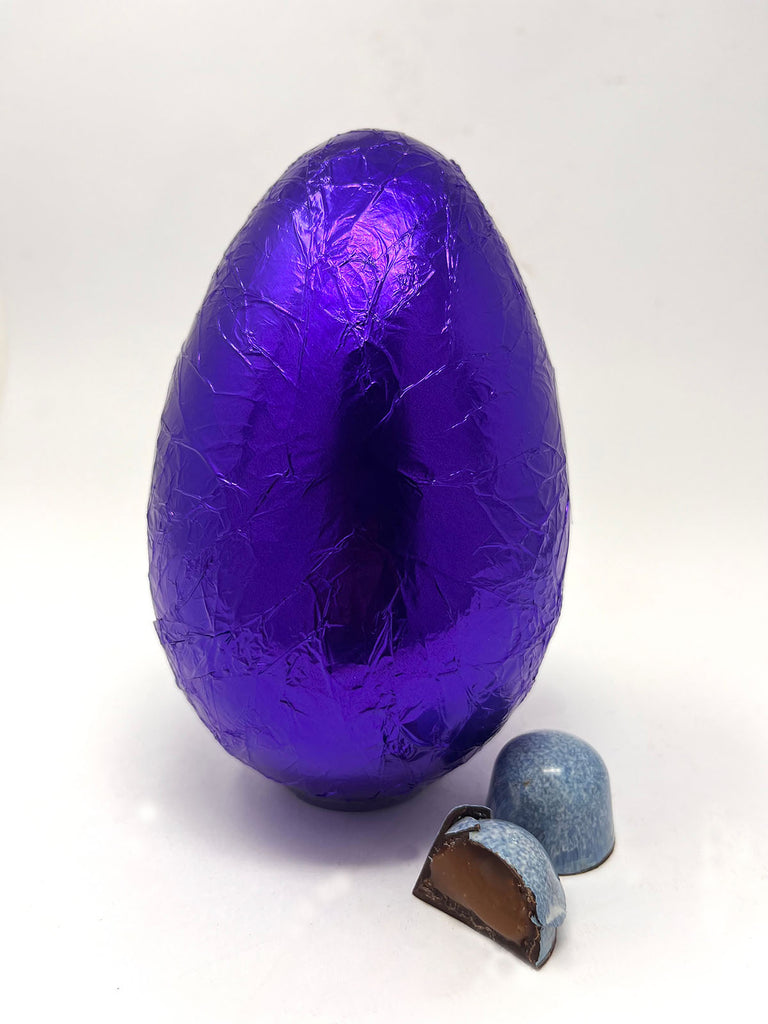 Dark Sea Salt Caramel Easter Egg in purple foil. Two Sea Salt caramel chocolates in the foreground. White background.