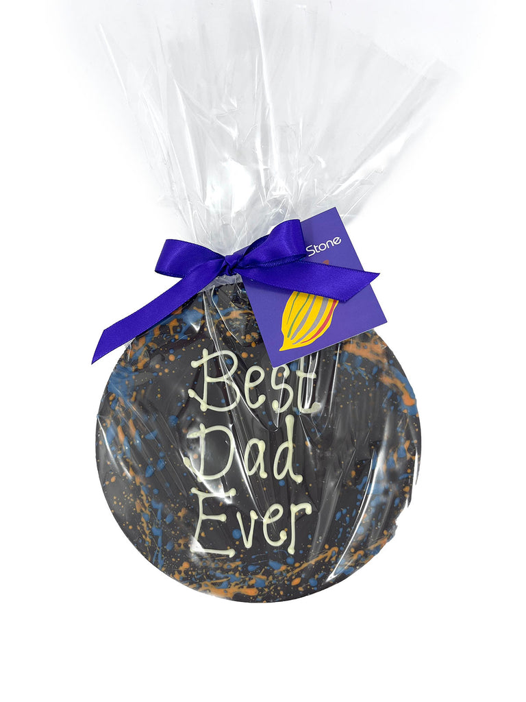 Round dark chocolate plaque decorated with orange and blue splashes and piped Best Dad Ever! Wrapped in cellophane and tied with a purple satin ribbon and gift tag. On a white background.