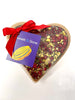 Dark Chocolate Heart decorated with scattered freeze-dried raspberries and crushed pistachios. Packaged in a clear heart shaped box with a ribbon and tag. On a white background.