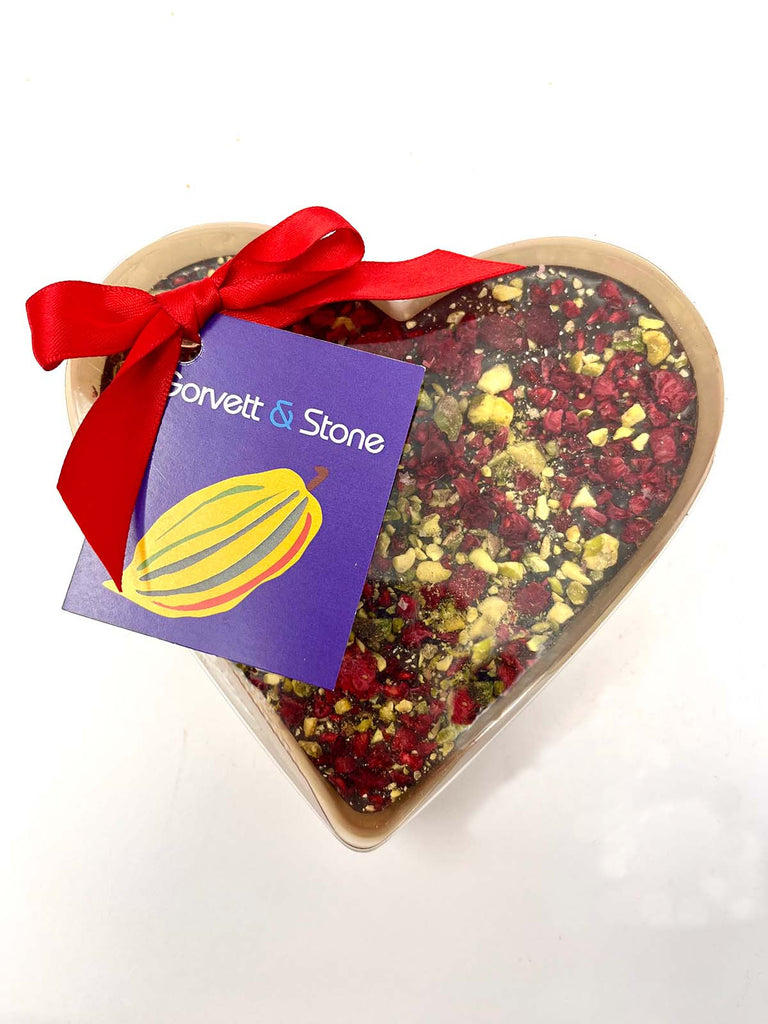 Dark Chocolate Heart decorated with scattered freeze-dried raspberries and crushed pistachios. Packaged in a clear heart shaped box with a ribbon and tag. On a white background.
