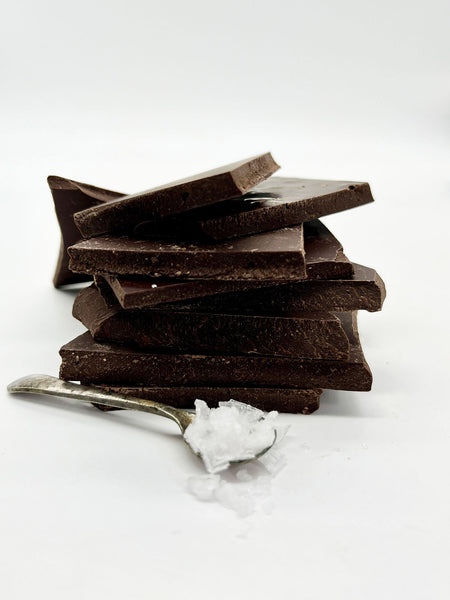 Stack of dark chocolate sea salt shards with a spoon of sea salt in front.  Taken on a white background.