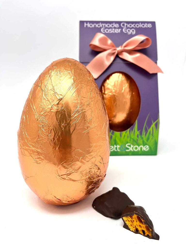 Dark Chocolate Easter Egg in copper foil with a boxed easter egg in the background and two pieces of dark chocolate cinder toffee in the foreground.  On a white background.