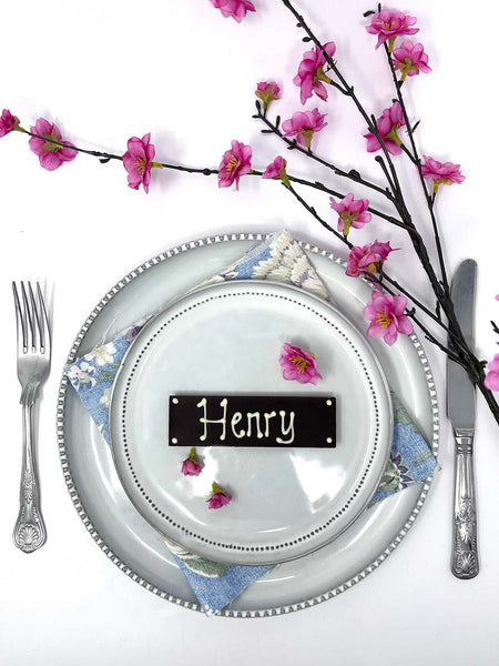 Decorative place setting with a dark chocolate bar as a name card labeled 'Henry', silverware, and cherry blossom branches on a white background.