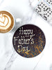One round dark chocolate father's day plaque. Decorated with blue and orange splashes and piped Happy Father's Day. On a white background marble backround with a cup of coffee and a newspaper crosswork in the background.