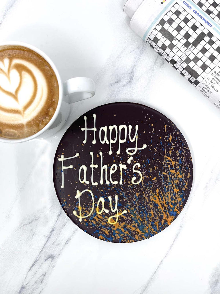 One round dark chocolate father's day plaque. Decorated with blue and orange splashes and piped Happy Father's Day. On a white background marble backround with a cup of coffee and a newspaper crosswork in the background.