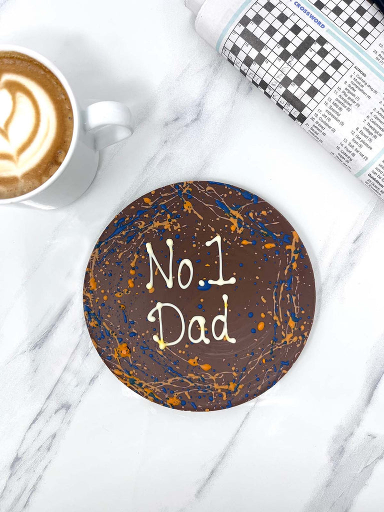 One round milk chocolate father's day plaque. Decorated with blue and orange splashes and piped No 1 Dad. On a white background marble backround with a cup of coffee and a newspaper crosswork in the background.