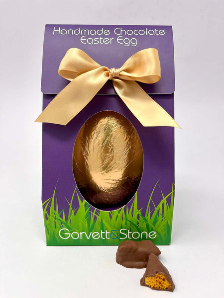 Gold foiled Easter Egg in purple Gorvett and Stone Box tied with a gold ribbon.  On a white background with 2 pieces of Cinder Toffee in the foreground.