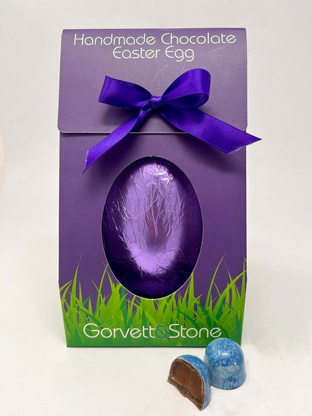 Milk Sea Salt Caramel Easter Egg in Gorvett & Stone Box and purple foil. Two Sea Salt caramel chocolates in the foreground. White background
