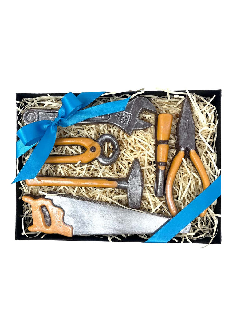 Realistic milk chocolate tools with red handles.  In a black gift box with cream shredded paper and a purple ribbon. On a white background.