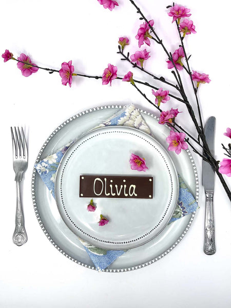 Decorative place setting with a milk chocolate bar as a name card labeled 'Henry', silverware, and cherry blossom branches on a white background.