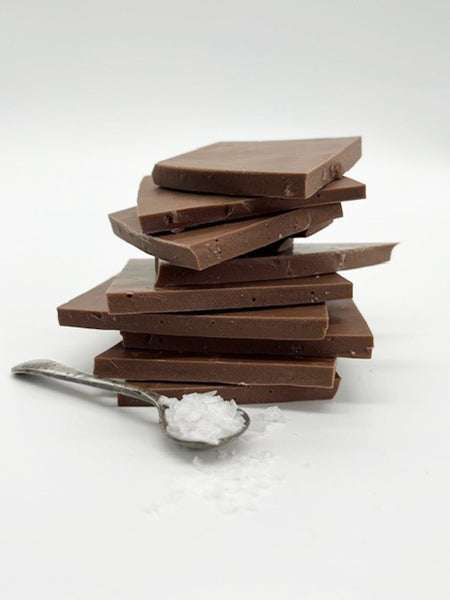 Stack of milk chocolate sea salt shards with a spoon of sea salt in front.  Taken on a white background.