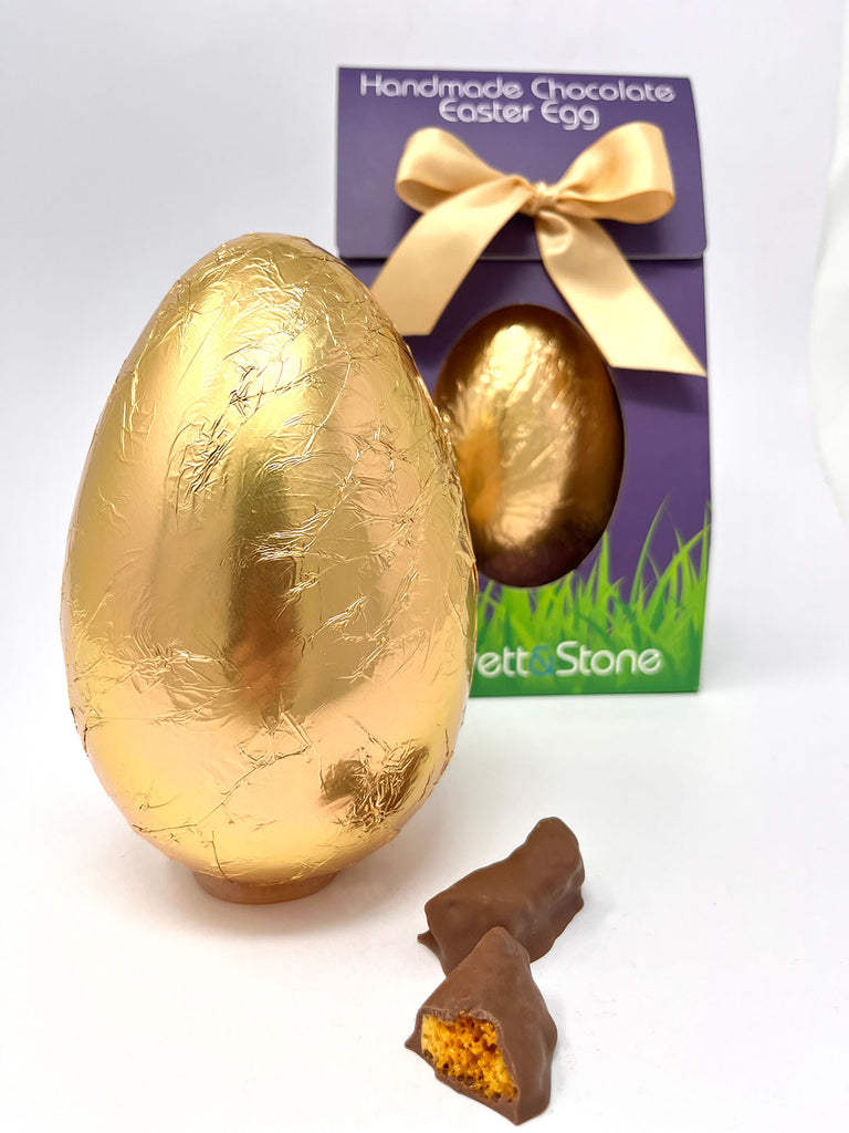 Milk Chocolate Easter Egg in goldr foil with a boxed easter egg in the background and two chunks of milk chocolate cinder toffee in the foreground.  On a white background.