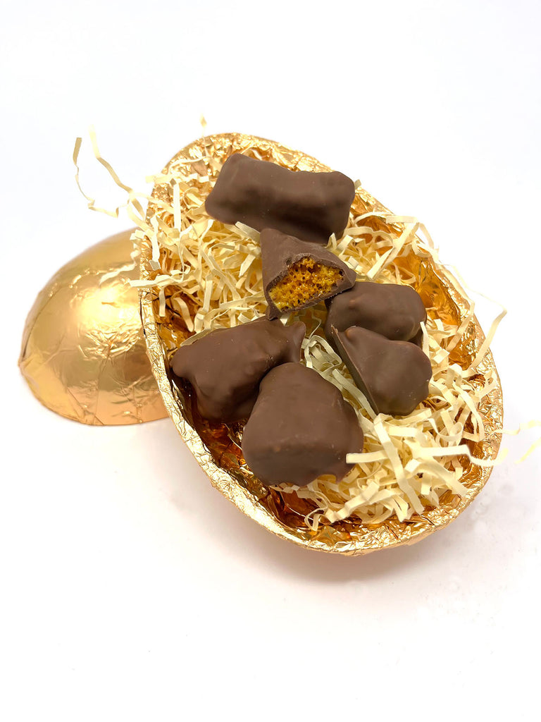 Open milk Chocolate Easter Egg in gold foil filled with pieces of milk chocolate cinder toffee on cream shredded paper.  On a white background.