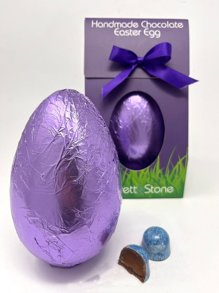 Milk Sea Salt Caramel Easter Egg in purple foil. Gorvett & Stone box in the background and  two Sea Salt Caramel chocolates in the foreground. White background.
