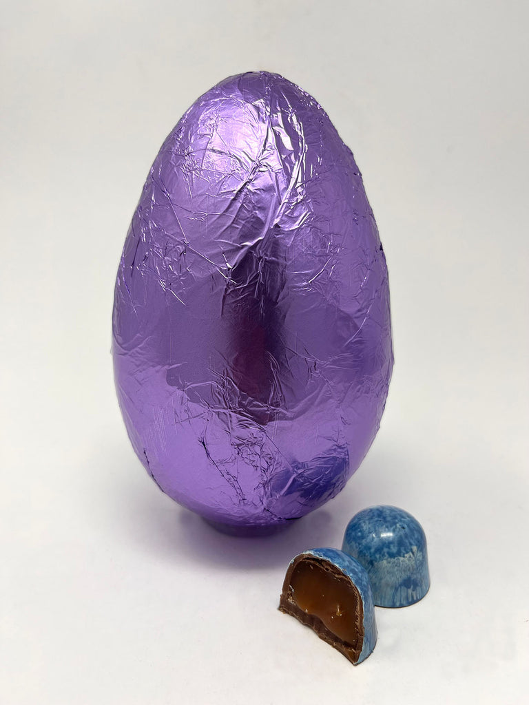 Milk Sea Salt Caramel Easter Egg in purple foil. Two Sea Salt caramel chocolates in the foreground. White background.