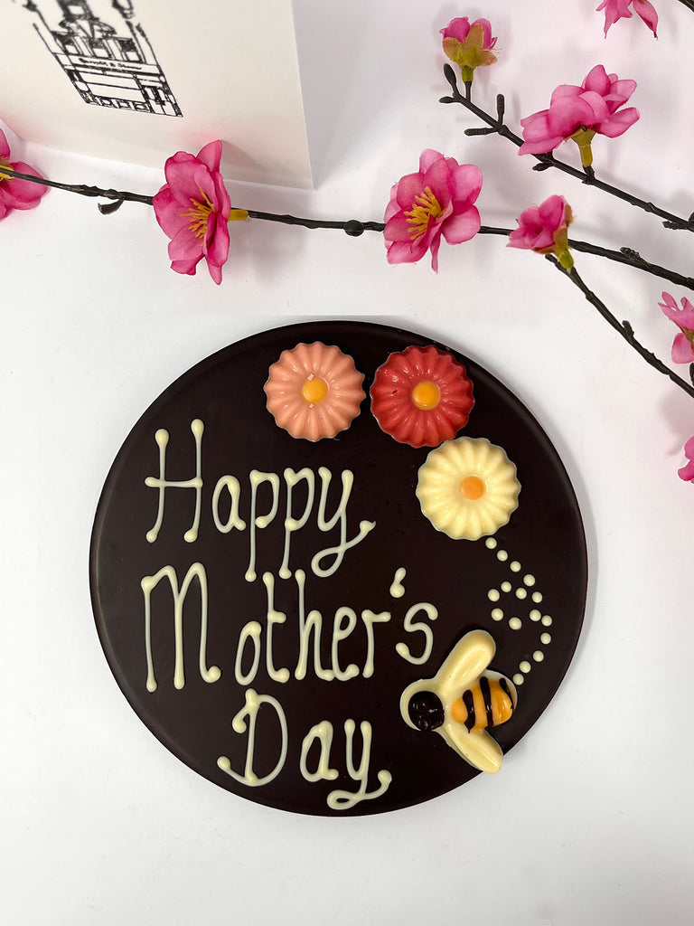 Dark chocolate plaque decorated with Happy Mother's Day, colourful chocolate flowers and a bumble bee. On a white background with pink flowers and a gift card in the background.