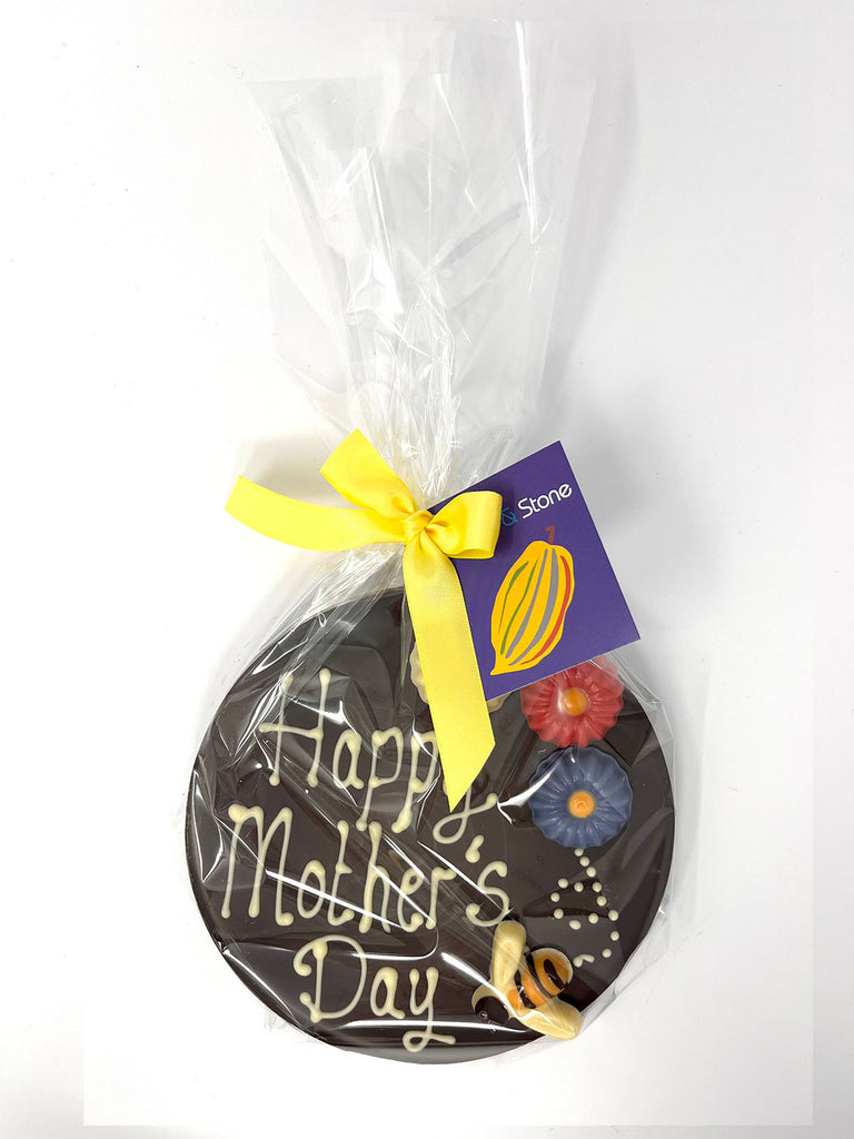 Dark chocolate plaque decorated with Happy Mother's Day, colourful chocolate flowers and a bumble bee. Wrapped in cellophane and tied with a satin ribbon and gift tag. On a white background with pink flowers and a gift card in the background.
