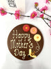 Milk chocolate plaque decorated with Happy Mother's Day, colourful chocolate flowers and a bumble bee. On a white background with pink flowers and a gift card in the background.