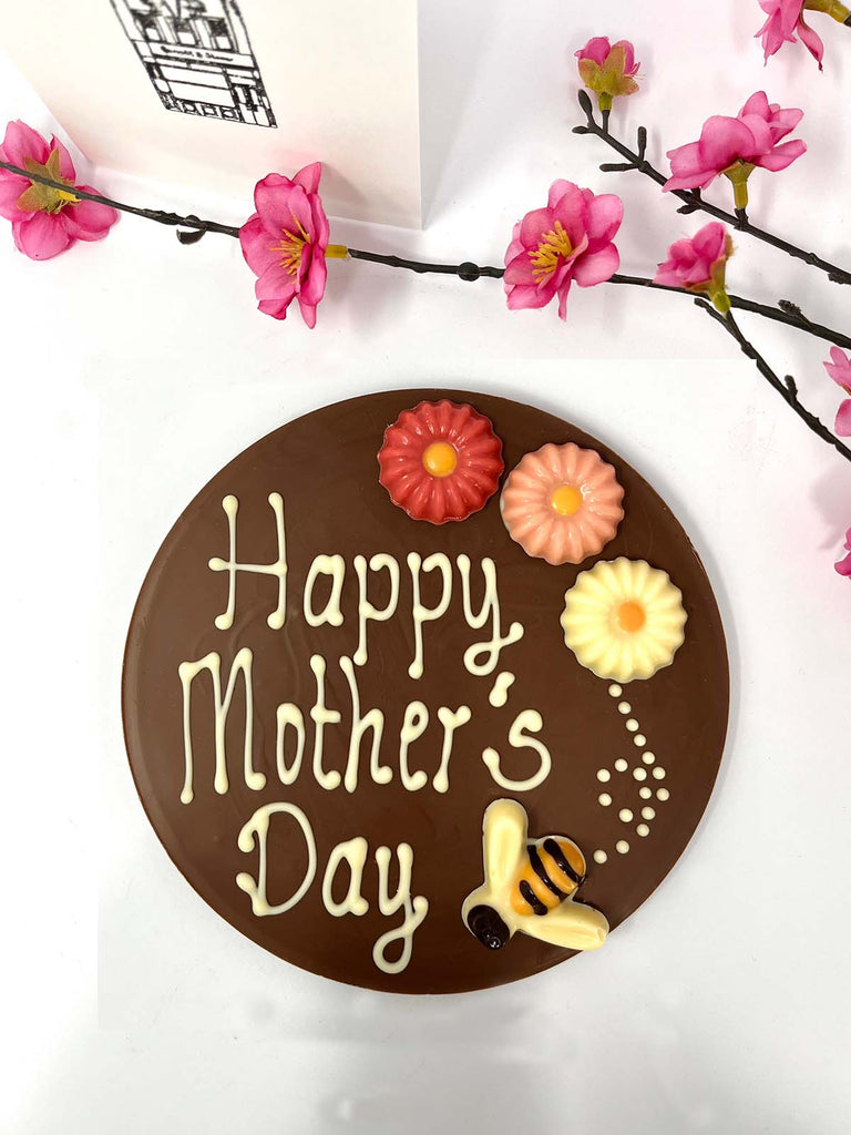 Milk chocolate plaque decorated with Happy Mother's Day, colourful chocolate flowers and a bumble bee. On a white background with pink flowers and a gift card in the background.