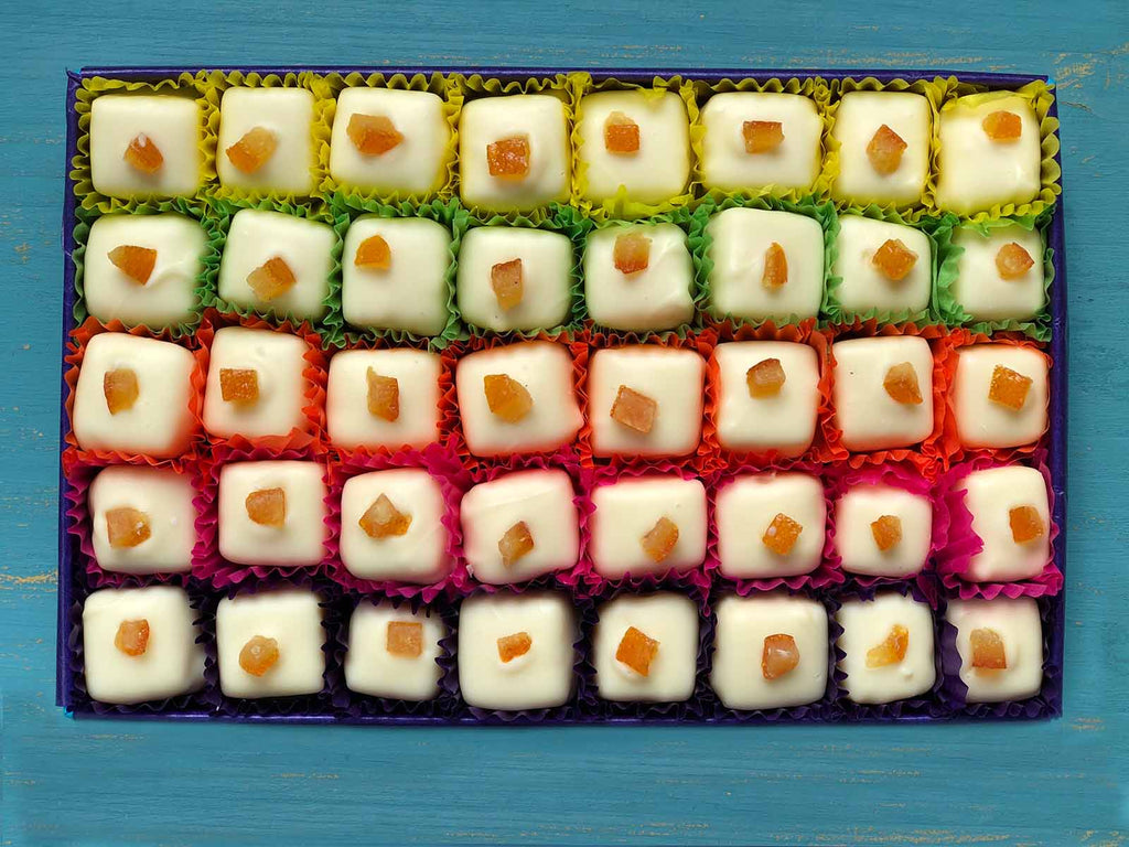 Open gift box of 40 white chocolates decorated with candied orange pieces. Each chocolate is contained in a colourful petits fours case. View from above on a painted blue background.