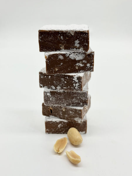 Stack of square peanut butter meltaways dusted with icing sugar. 3 peanuts scattered in front of meltaways. On a white background. 