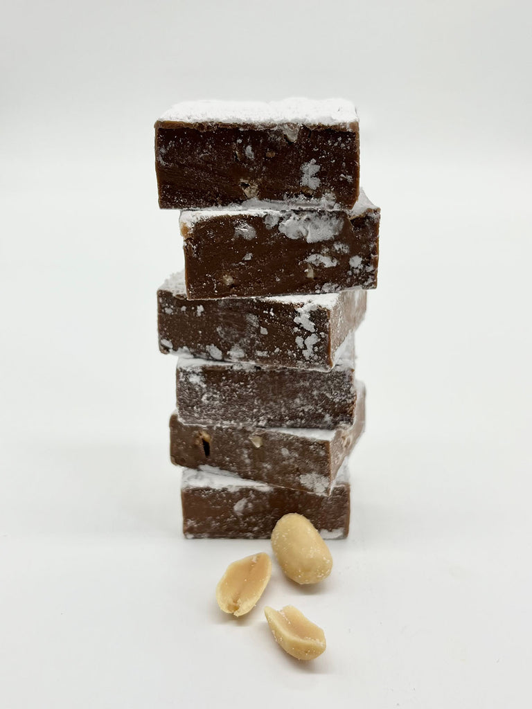 Stack of square peanut butter meltaways dusted with icing sugar. 3 peanuts scattered in front of meltaways. On a white background. 