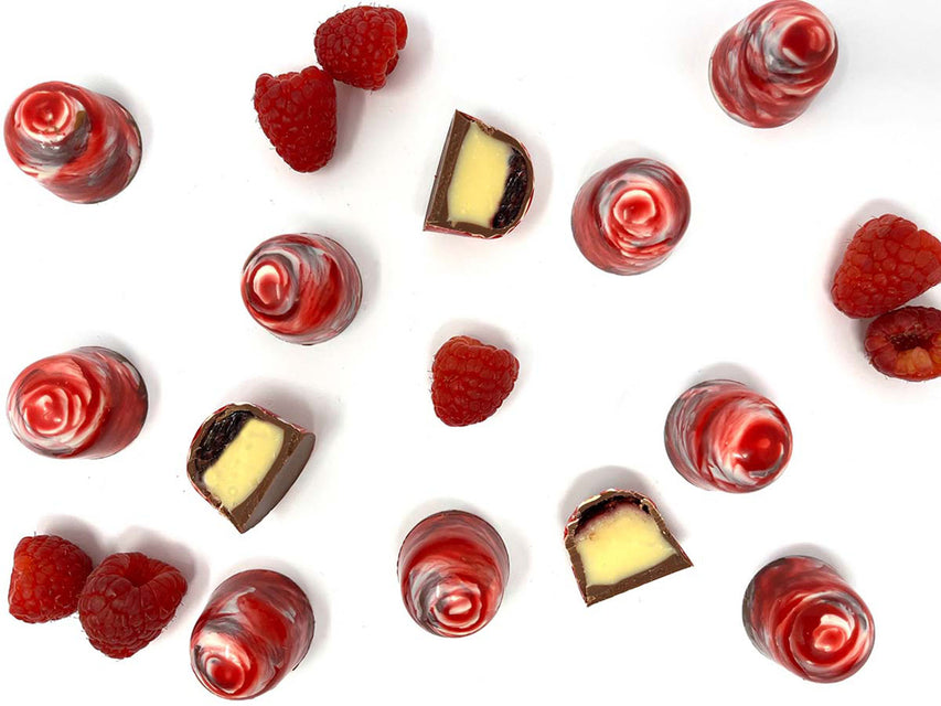 Raspberry Cream Tea Chocolates decorated with red and white shiny cocoa butter swirls, and fresh raspberries scattered on a white background. Three chocolates are cut open to show the layers of jam and ganache inside. The image is taken from above. 