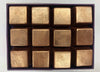Twelve square metallic gold chocolates arranged in a 3x4 grid inside a dark purple box.