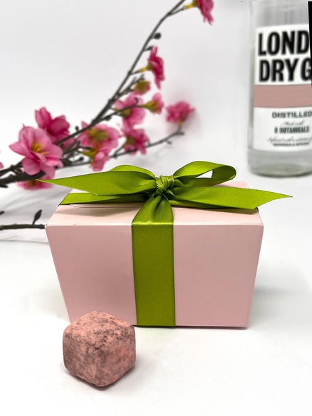 Pink gift box with a green ribbon, a pink chocolate truffle, and a bottle of gin on a white background with pink flowers.