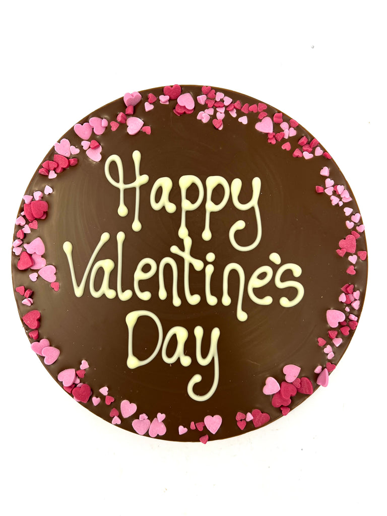 Milk chocolate round plaque on a white background. Plaque is piped with 'Happy Valentine's Day,  with a border of pink and red sugar hearts.