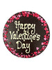 Dark chocolate round plaque on a white background. Plaque is piped with 'Happy Valentine's Day,  with a border of pink and red sugar hearts.