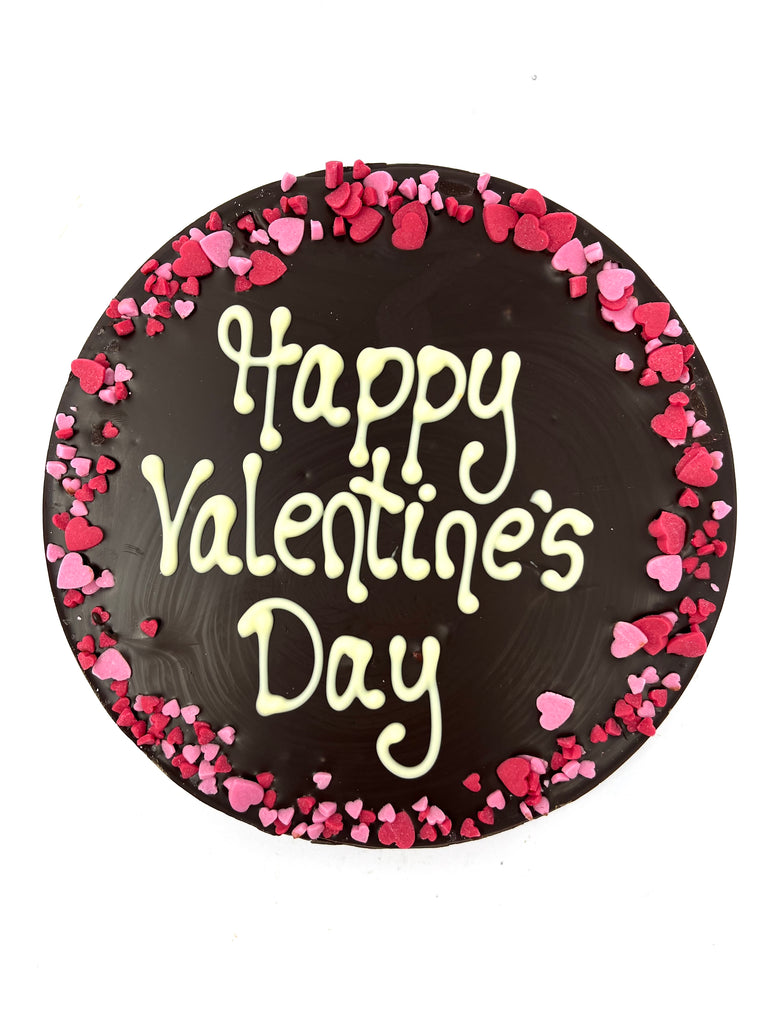 Dark chocolate round plaque on a white background. Plaque is piped with 'Happy Valentine's Day,  with a border of pink and red sugar hearts.