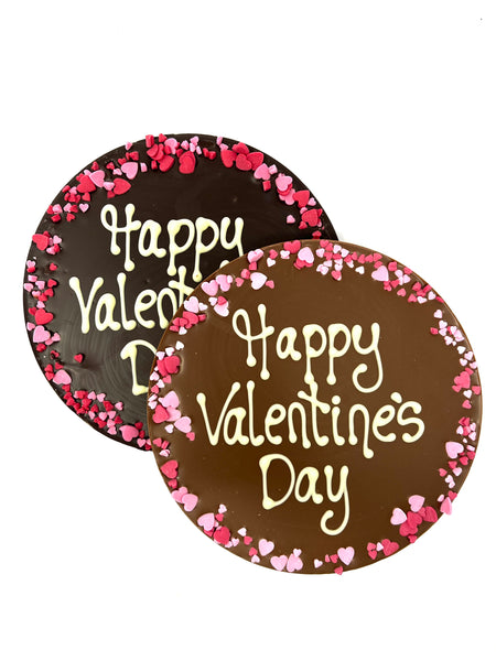 Two round chocolate plaques on a white background. Plaques are piped with 'Happy Valentine's Day,  with a border of pink and red sugar hearts.