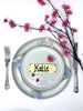 Decorative place setting with a white chocolate bar as a name card labeled 'Henry', silverware, and cherry blossom branches on a white background.